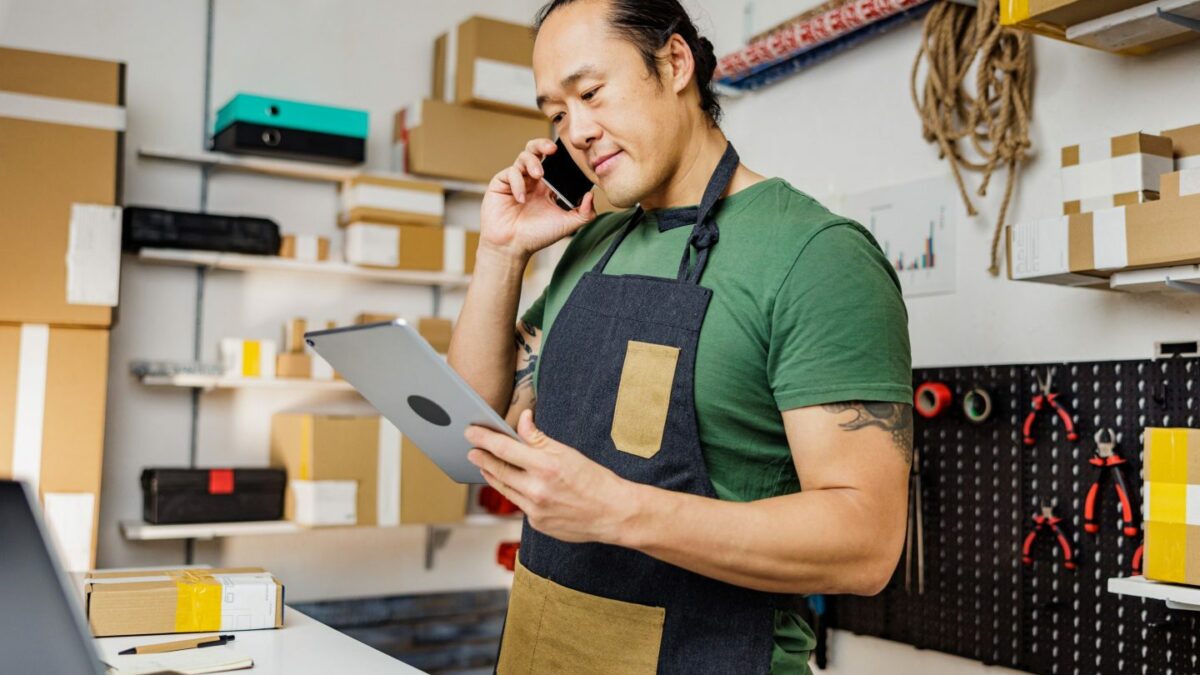 A person wearing an apron in a storeroom, talking on the phone looking at an iPad.