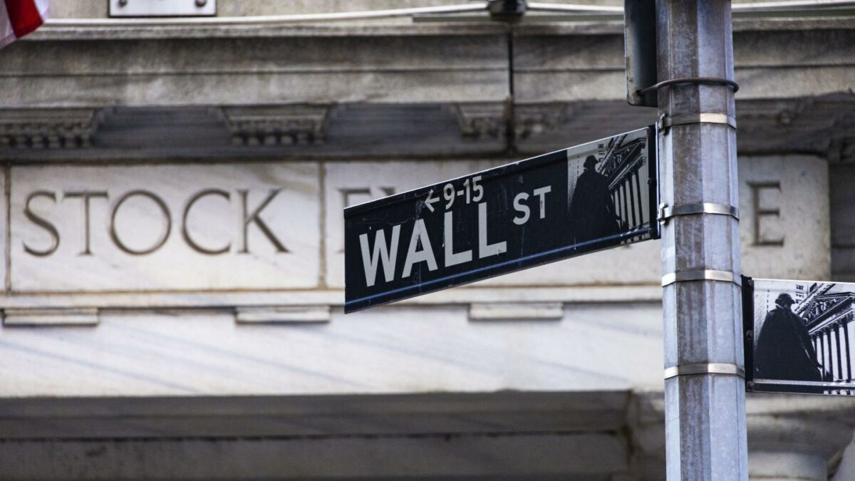 Wall Street sign inscription as seen on the road and buildings of Wall St with the entrance of New York Stock Exchange in the background.